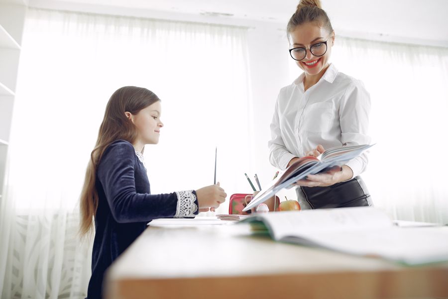 A tutor smiling as she goes through a workbook with a young student at a bright desk.